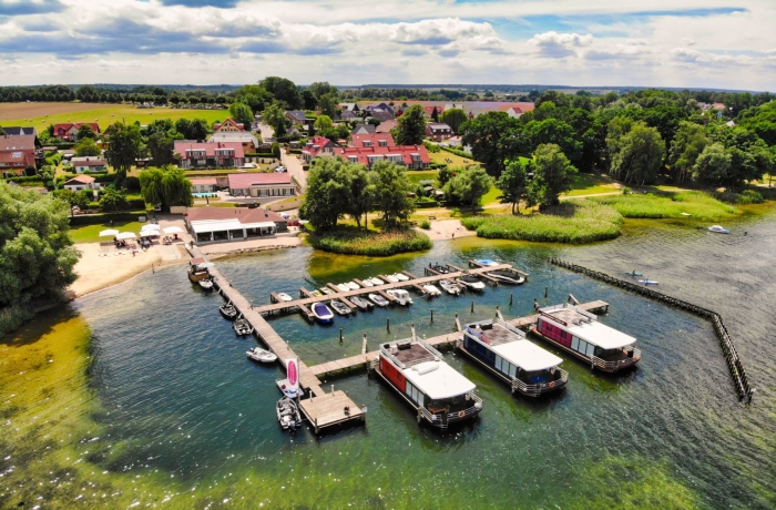 Luftaufnahme eines idyllischen Resorts am See mit kleinem Yachthafen, Hausbooten, Sandstrand und grüner Landschaft unter weitem Himmel.