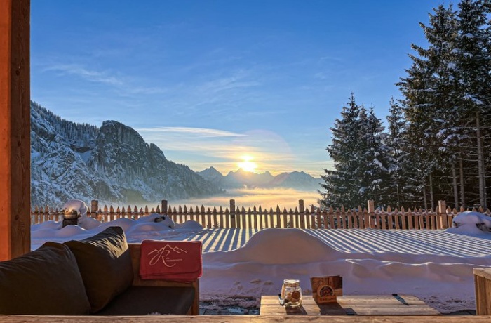 "Eine atemberaubende Aussicht vom Hotel Das Rübezahl - Romantic Hideaway & Boutique Spa auf eine verschneite Berglandschaft bei Sonnenaufgang. Im Vordergrund steht eine gemütliche Sitzgelegenheit auf einer Terrasse, umgeben von Schnee, mit einem Tisch, auf dem Getränke und Snacks stehen. Die Sonne geht über den Gipfeln auf, während der Himmel in klarem Blau erstrahlt und Nebel sanft die Täler füllt. Tannenbäume und ein hölzerner Zaun rahmen die Szene malerisch ein.