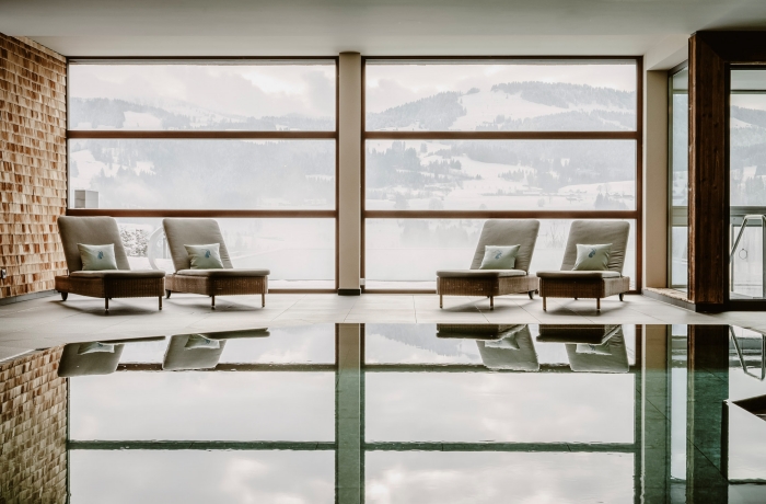 Ruhiger Indoor-Pool mit gemütlichen Liegen vor großen Panoramafenstern und verschneiter Berglandschaft im Hintergrund.
