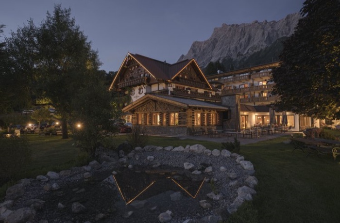 Abendansicht des Hotel Spielmann mit traditioneller alpenländischer Architektur, beleuchtet, umgeben von einem gepflegten Garten und einem kleinen Teich, vor der Kulisse eines majestätischen Bergmassivs