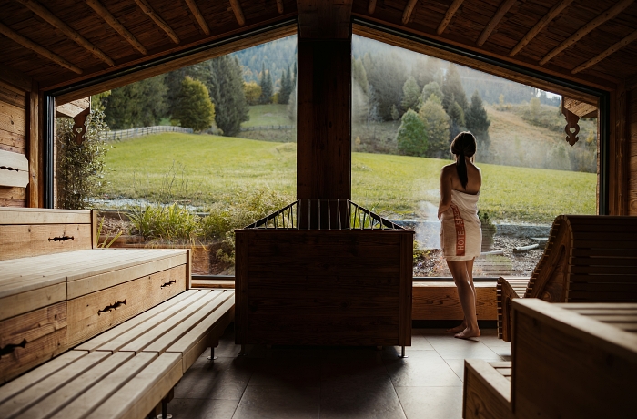 Eine Frau im Saunatuch schaut durch die Panoramafenster des Hotel Spielmann auf grüne Almwiesen und Tannenbäume