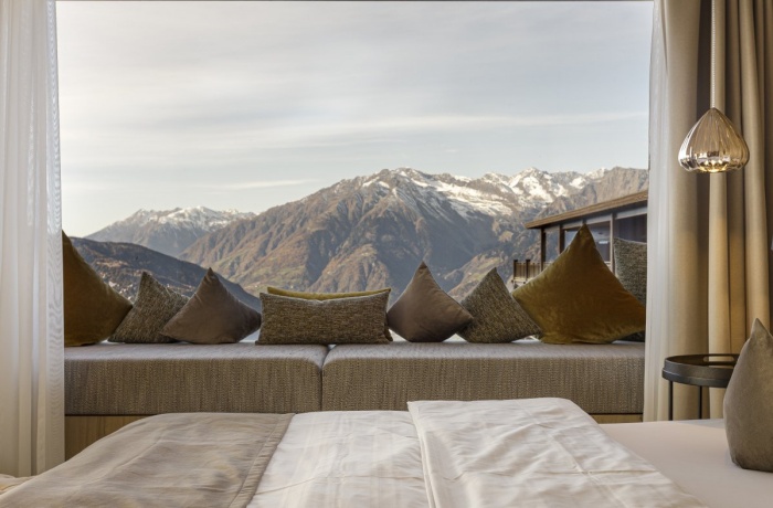 Gemütliches Hotelzimmer im Hotel Chalet Mirabell mit großem Panoramafenster, das einen Blick auf schneebedeckte Berge und die umliegende Natur bietet, dekoriert mit weichen Kissen auf einer Fensterbank.