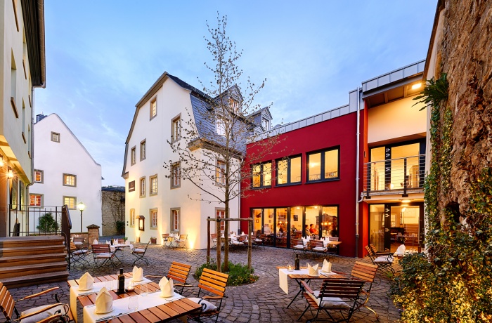 Stimmungsvoller Innenhof eines Restaurants oder Hotels mit gedeckten Holztischen, umgeben von historischen und modernen Gebäuden in warmem Abendlicht.