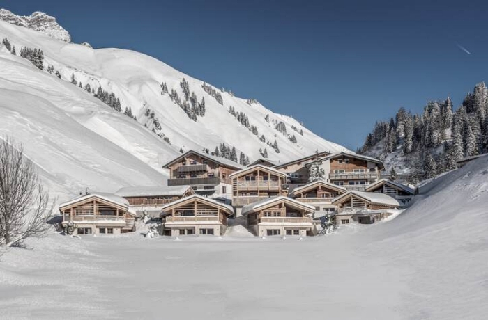 Ein Chalet-Dorf liegt in einem verschneiten, kleinen Tal. Zur linken Bildseite erstrecken sich aufwärts verschneite Berge, zur rechten Bildseite ein verschneiter Tannenwald.