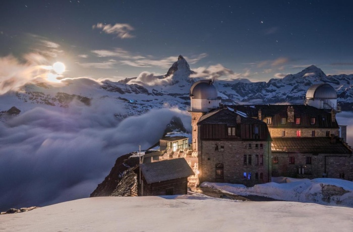 Das 3100 Kulmhotel Gornergrat bei Nacht, umgeben von schneebedeckten Bergen und einem wolkenverhangenen Tal, mit Blick auf das Matterhorn und einen klaren Sternenhimmel.