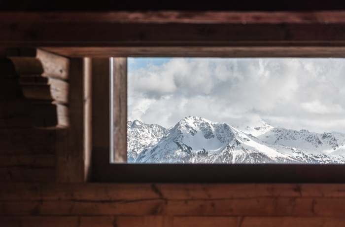 Blick durch ein Holzfenster im LeCrans Hotel auf schneebedeckte Berge und bewölkten Himmel, eingerahmt von rustikalen Holzbalken.