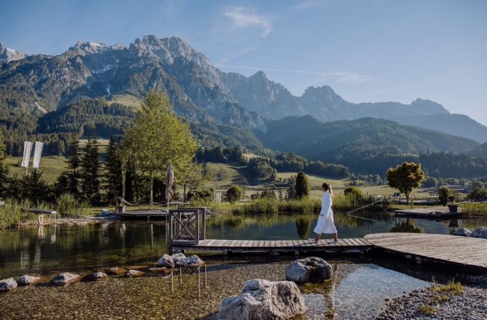 Person im Bademantel spaziert entlang eines Steges über einen Naturteich im Naturresort Puradies, eingebettet in eine malerische Alpenlandschaft mit imposanten Bergen im Hintergrund.