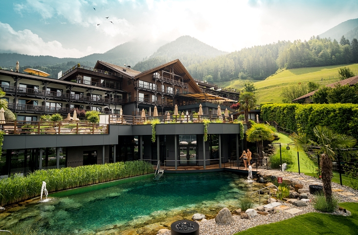 Blick auf das hundefreundliche Hotel Andreus Resorts in Südtirol, im Hintergrund Berge und Wälder, das Hotelgebäude mit Wellnessbereich im Erdgeschoss und Naturpool