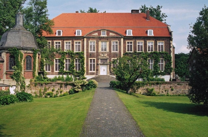 Historisches Herrenhaus mit rotem Dach und efeubewachsenen Fassaden, erreichbar über einen gepflasterten Weg durch eine gepflegte Gartenanlage.