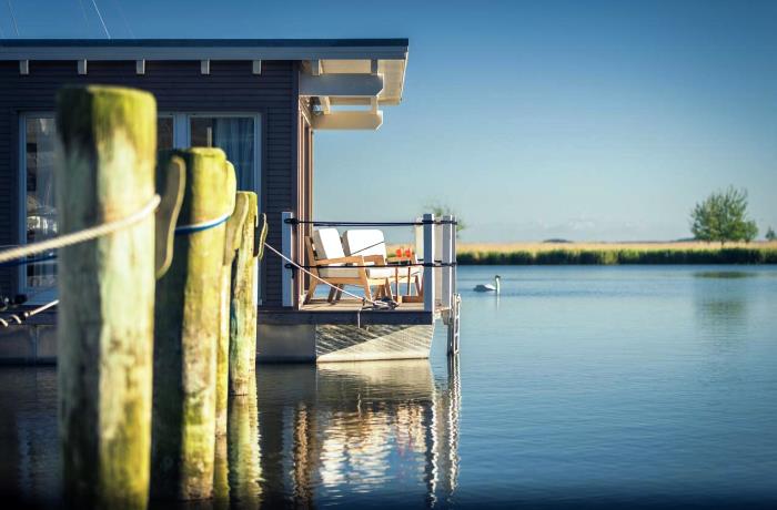 Hotels an der Nordsee und Ostsee: Ein Hausboot mit bestuhlter Terrasse liegt auf ruhiger Wasseroberfläche.