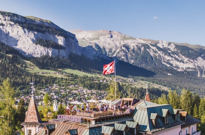 Über der Dachterrasse des Hotelgebäudes ist die Schweizer Flagge gehisst – im Hintergrund blickt man auf die Berge