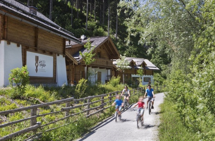 Eine vierköpfige Familie fährt auf Fahrrädern am bergdorfartig-gestaltetem Hotel Berglebn vorbei