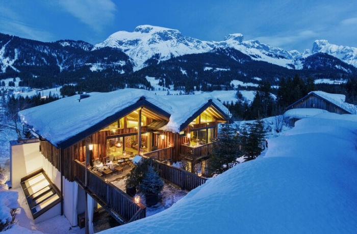 Skiurlaub: In der Abenddämmerung sind zwei verschneite Chalets mit großer Fenstefront zu sehen, im Inneren brennt warmes Licht.