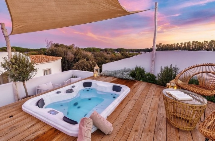 Abendlicher Blick auf das Hotel Corte Bianca auf Sardinien mit einem malerischen lila-rosa Abendhimmel im Hintergrund. Ein Sonnensegel spannt sich über einer hölzernen Terrasse, auf der ein luxuriöser Whirlpool eingelassen ist. Die idyllische Szenerie bietet Entspannung und Eleganz für einen unvergesslichen Aufenthalt