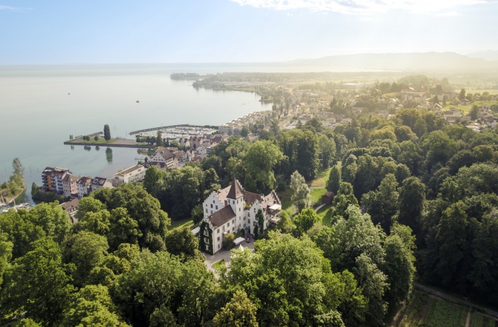 Luftaufnahme des Schloss Wartegg, umgeben von dichtem, grünem Wald. Im Hintergrund liegt ein kleiner Ort mit Häusern und einem Yachthafen am Ufer des Bodensees