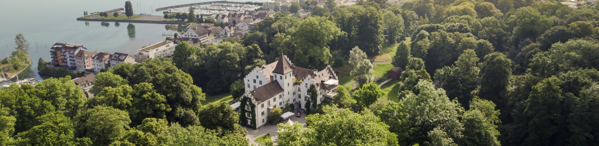 Luftaufnahme des Schloss Wartegg, umgeben von dichtem, grünem Wald. Im Hintergrund liegt ein kleiner Ort mit Häusern und einem Yachthafen am Ufer des Bodensees.