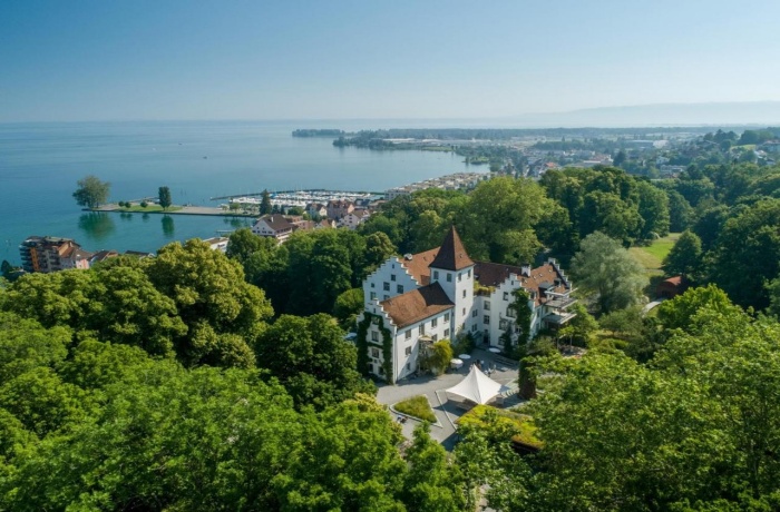 Luftaufnahme von Schloss Wartegg mit roten Dächern, umgeben von Bäumen, mit Blick auf den Bodensee und den Hafen von Rorschach.