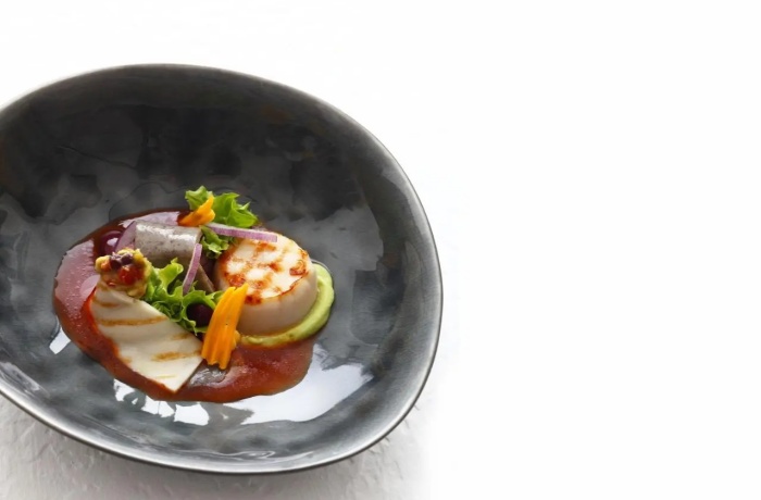 Hotels mit Gourmetrestaurant: Modern angerichtetes Gourmetgericht in schwarzer Schale: Gegrillte Jakobsmuschel mit Avocadocreme, Tomatensauce, Salat und essbaren Blüten.