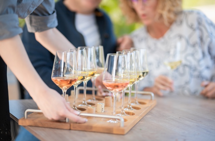Mehrere gefüllte Weingläser in einem Holzträger auf einem Tisch, während Personen im Hintergrund Wein verkosten.