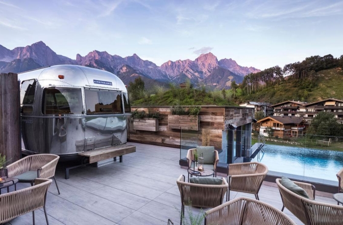 Terrasse mit Sitzbereich und umgebautem Wohnwagen vor beeindruckender Bergkulisse und entspannter Urlaubsatmosphäre.

