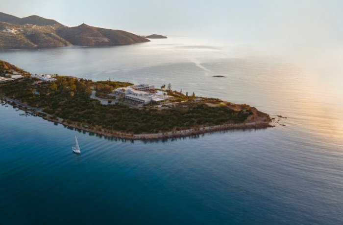 Eine Halbinsel auf Kreta, umgeben von ruhigem, blauem Meer, mit Gebäuden und üppiger Vegetation. Im Vordergrund segelt ein kleines Boot durch das Wasser, während sich im Hintergrund die Küstenlinie und Hügel der Insel abzeichnen.