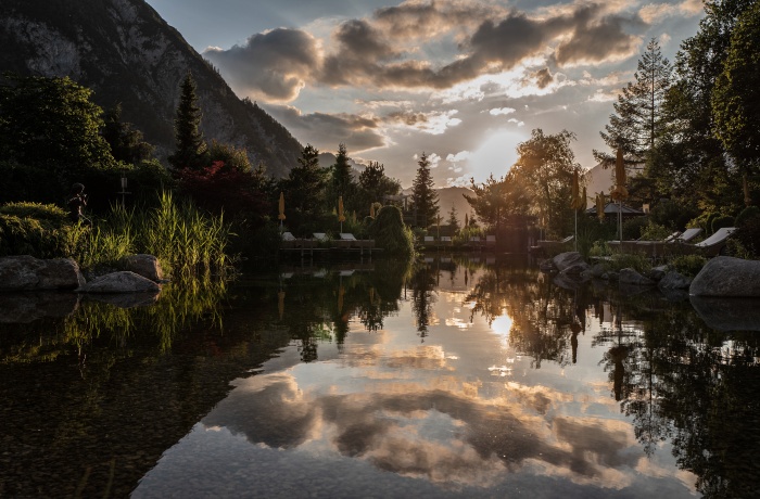 Abendstimmung in der Wellnessresidenz Alpenrose in Maurach: Die Sonne geht über dem Karwendelgebirge unter, ihr warmes Licht spiegelt sich im klaren Naturteich des Hotelgartens. Rundherum liegen Liegen, Schilf und Bäume in goldenes Licht getaucht – eine friedliche Szene zwischen Bergen und Wasser.