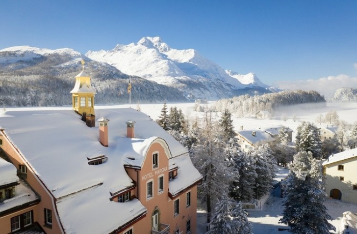 Im Vordergrund ist das Hotel, es hat eine Orange-braune Fassade und einen kleinen goldenen Turm. Im Hintergrund  sieht man einen großen Schneebedeckten Berg und viele Schneebedeckte Bäume.