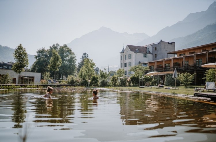 Reisetrends 2024: Zwei Frauen schwimmen im Naturteich des VillaVerde Aparthotel, im Hintergrund sieht man die Südtiroler Berglandschaft