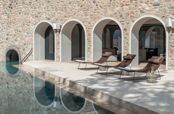 Mediterraner Poolbereich mit drei braunen Sonnenliegen und Beistelltisch auf einer hellen Steinterrasse vor einer Steinmauer mit eleganten Rundbögen. Der Pool reflektiert die Architektur, eine kleine Tunnelöffnung führt vom Wasser in das Gebäudeinnere.