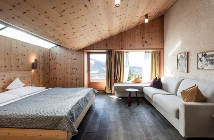 Zimmer mit einer Decke aus Holz und Wänden aus Holz. Ein großes Bett und ein Sofa. Die Fenster sind ausgerichtet in Richtung Berge, es liegt leichter Schnee.