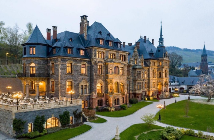 Prächtiges Schloss im neogotischen Stil mit Türmen und verzierten Fassaden, abends warm beleuchtet, mit Terrasse und Blick auf gepflegte Gartenanlage.