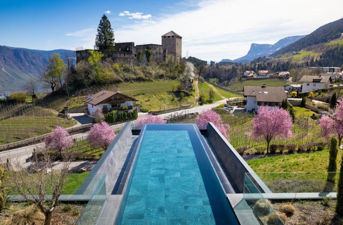 Blick aus dem Infinitypool auf rosa Kirschblüten und im weiteren Hintergrund auf eine Burganlage und die Südtiroler Berge