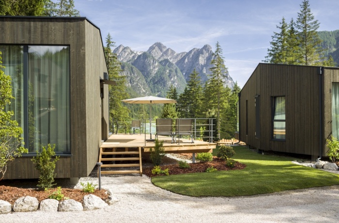 Moderne Skyview Chalets am Toblacher See, umgeben von Bergen und Wald, mit Terrasse und Sonnenschirm, eingebettet in eine ruhige Naturlandschaft.
