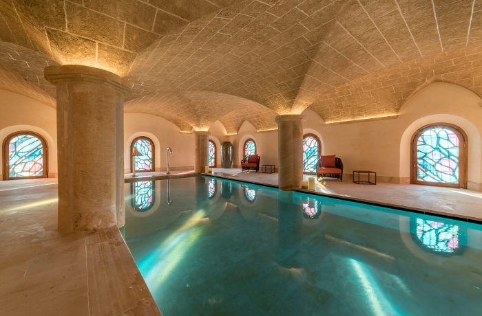 Eleganter Indoor-Pool in einem historischen Gewölbesaal mit sandfarbenen Steinbögen, dicken Säulen und kunstvoll gestalteten Buntglasfenstern. Warmes Licht betont die Architektur, Liegen an den Seiten laden zum Entspannen ein.