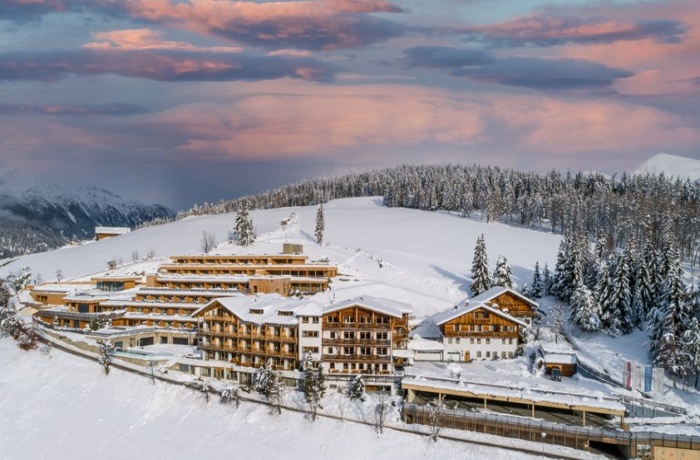 Im Fokus des Bildes sind die Gebäude der Hotel anlege, welche im Chaletstil erbaut sind, im Hintergrund ist ein Sonnenuntergang und Schneebedeckte  Tannen