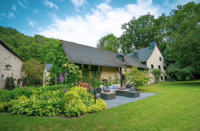 Helles Landhaus mit schrägem Dach und überdachter Terrasse, Sitzmöbeln und üppigem Garten mit Blumen und Sträuchern.