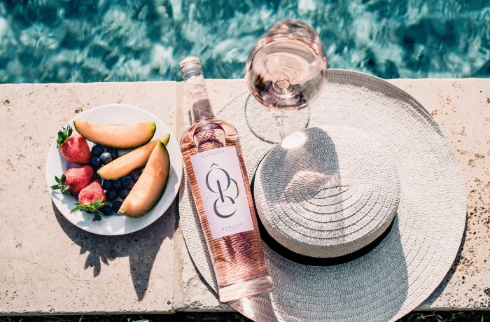 Hotels mit Gourmetrestaurant: Sommerliche Szene am Pool mit Roséweinflasche, gefülltem Weinglas auf einem Strohhut und einem Teller mit Melone, Erdbeeren und Blaubeeren.