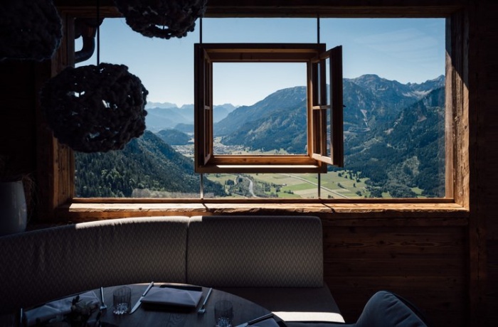 Blick aus einem Fenster im Hotel Blaue Burg auf eine weitläufige Berglandschaft. Durch das offene Fenster sieht man grüne Täler und bewaldete Berghänge unter einem klaren blauen Himmel. Im Vordergrund befinden sich gemütliche Sitzgelegenheiten in einem rustikalen Raum mit Holzwänden.