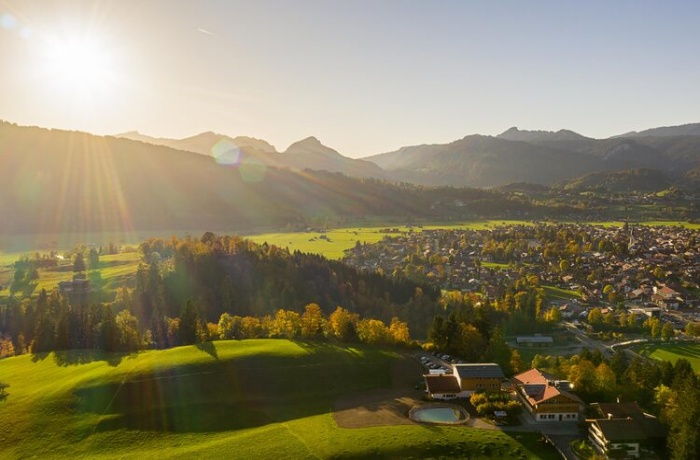 Brückentage 2025: Eine malerische Landschaft im Allgäu bei Sonnenuntergang oder Sonnenaufgang: grüne Hügel, weitläufige Wiesen, ein kleines Dorf eingebettet in das Tal, umgeben von den majestätischen Alpen. Die tiefstehende Sonne wirft warme Lichtstrahlen über die Szenerie und erzeugt eine friedliche Atmosphäre.