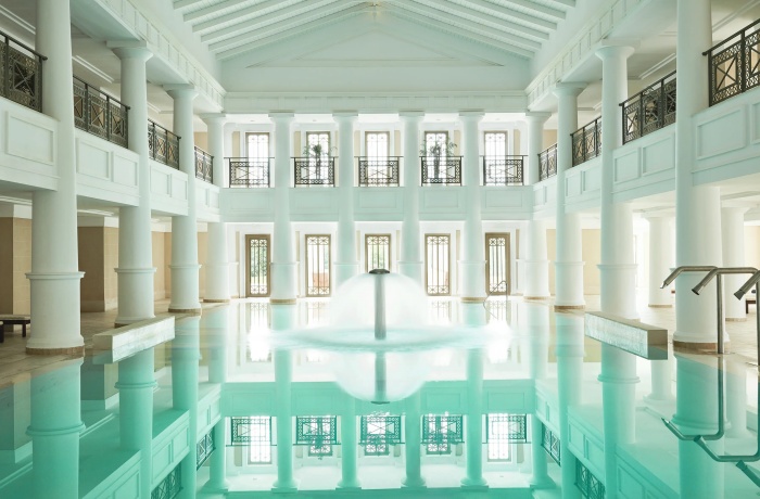 Prächtiger Innenpool im Stil eines antiken Atriums mit hohen weißen Säulen, zwei umlaufenden Galerien und großen Fenstern, die Tageslicht hereinlassen. In der Mitte des türkisfarbenen Beckens sprudelt ein symmetrischer Springbrunnen.