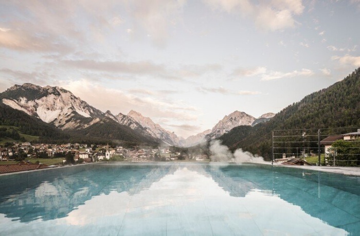 Infinity-Pool im Excelsior Dolomites Life Resort mit Blick auf ein idyllisches Dorf und die umgebenden Dolomitenberge bei Sonnenuntergang.