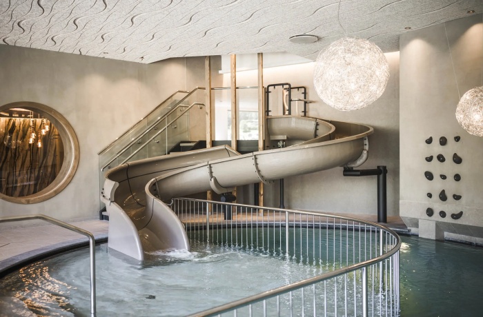 Innenbereich eines modernen Wasserparks mit einer grauen Röhrenrutsche, die in ein flaches Rundbecken mündet. Die Szene ist mit Designerlampen, gläsernem Treppenaufgang und einer kleinen Kletterwand stilvoll gestaltet.