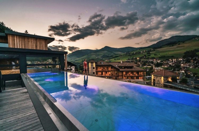 Eine Frau in rotem Badeanzug läuft auf dem Rand eines Infinity-Sky-Pools. Im Hintergrund sieht man einen Sonnenuntergang sowie sanfte Berge