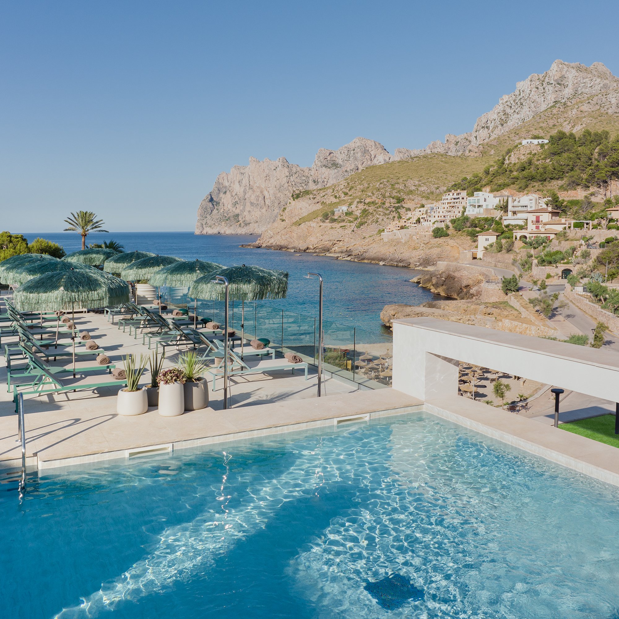 Traumhafte Panoramakulisse mit Infinity-Pool und Blick auf die malerische Küstenlandschaft. Sonnenliegen mit stilvollen Bastschirmen laden auf der großzügigen Terrasse zum Entspannen ein, während das kristallklare Wasser der Bucht im Sonnenlicht glitzert. Im Hintergrund erheben sich beeindruckende Felsen und eine charmante Küstensiedlung – ein perfektes Hideaway für Luxus und Naturgenuss.