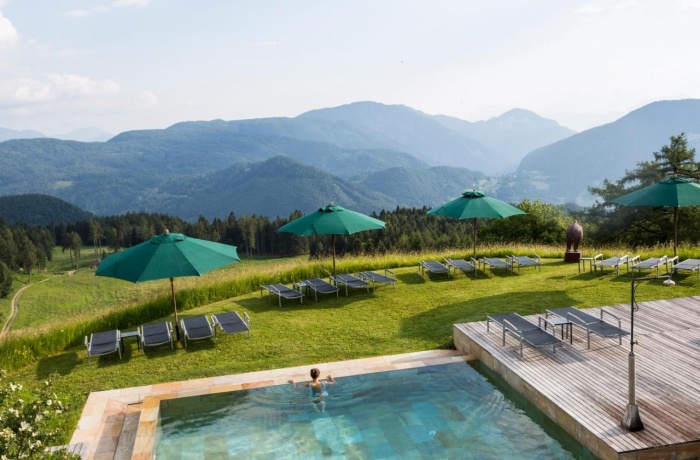Außenpool mit Sonnenliegen und grünen Sonnenschirmen, umgeben von Wiesen und Wäldern, mit Blick auf bewaldete Berge.