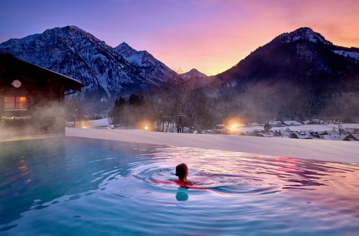 Person schwimmt im beheizten Außenpool des Hotel Prinz-Luitpold-Bad bei Sonnenuntergang, umgeben von schneebedeckten Bergen und einer malerischen Winterlandschaft.