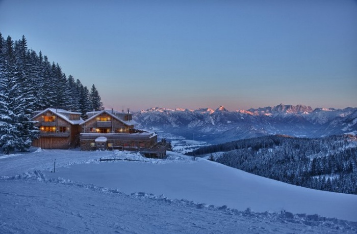 Winterlandschaft mit den schneebedeckten BöckLodges, umgeben von verschneiten Bäumen und mit Blick auf ein Bergpanorama bei Sonnenuntergang.