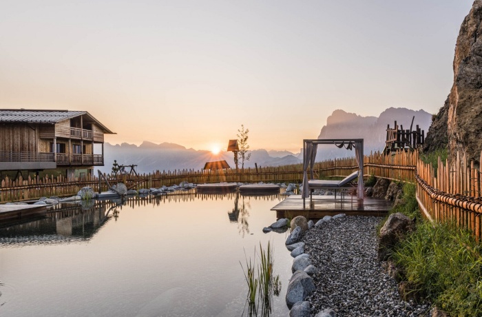 Naturteich bei Sonnenaufgang im Hotel Goldknopf, umgeben von Holzgebäuden und einer malerischen Berglandschaft, mit einer gemütlichen Liegefläche am Steg.