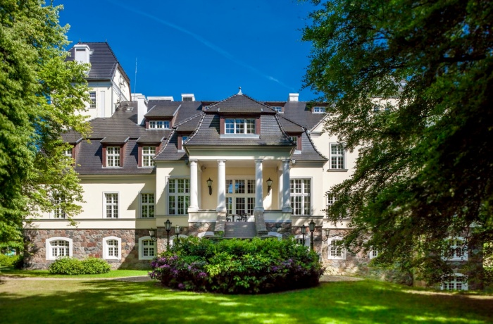 Elegantes Herrenhaus mit weißer Fassade, Säulenportal und dunklem Dach, umgeben von gepflegtem Park und grünen Bäumen bei sonnigem Wetter.