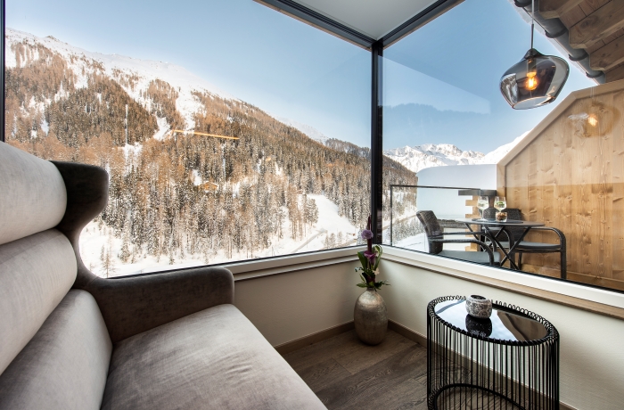 Gemütlicher Sitzbereich in einem Hotelzimmer mit großem Fenster und Ausblick auf eine verschneite Berglandschaft.